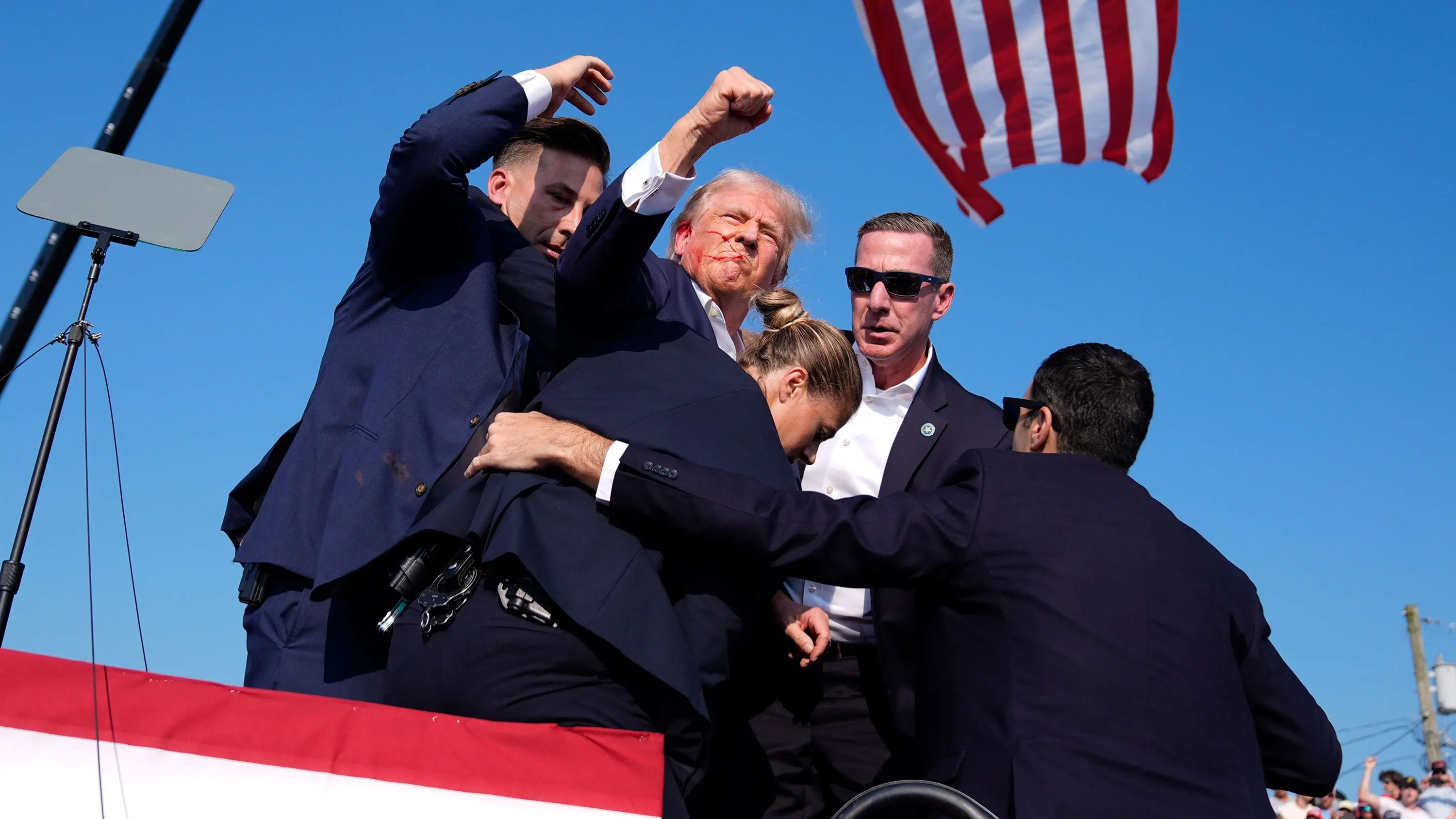 Security guards surround Donald Trump during a tense moment at a rally, U.S. flag in background.