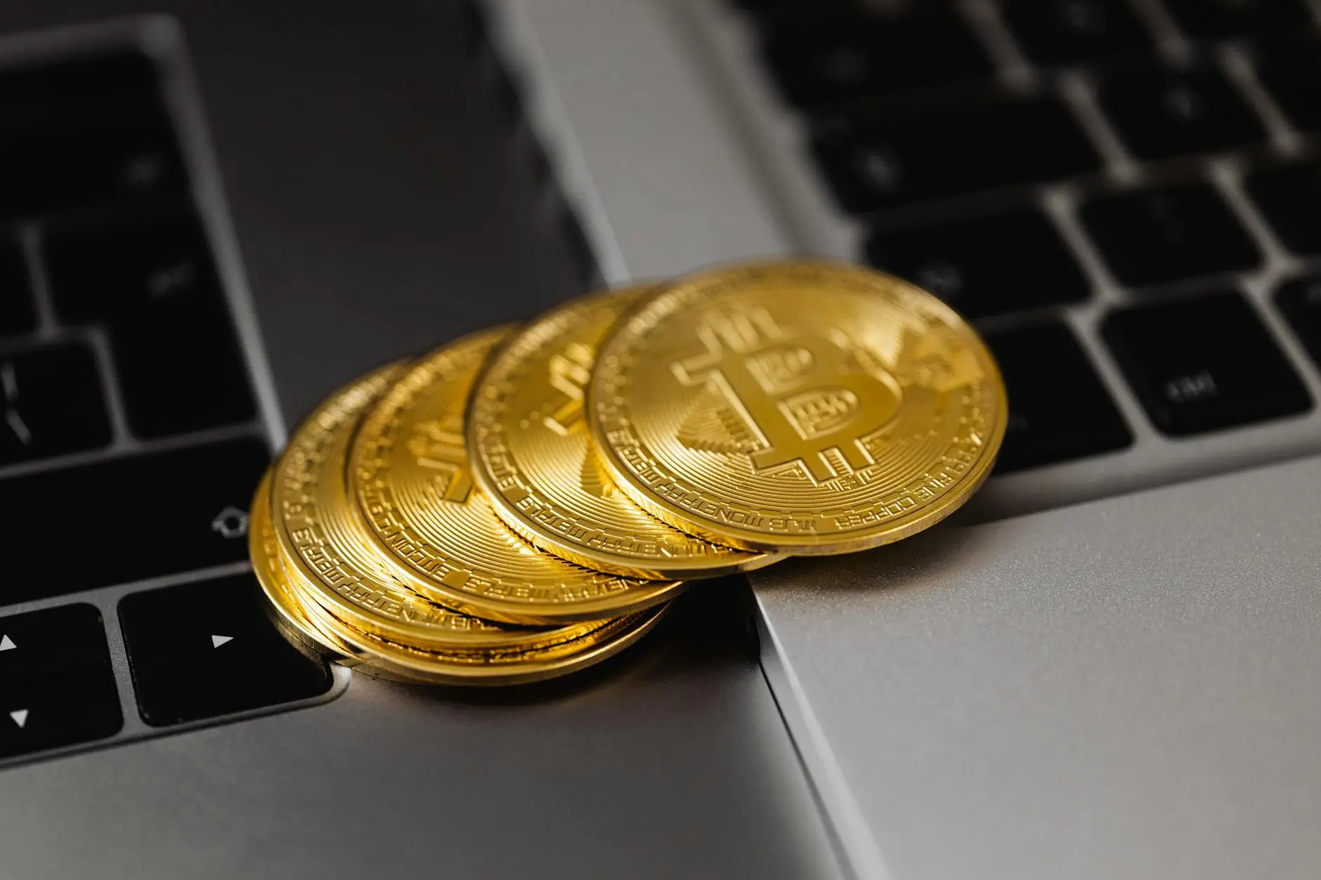 Four gold Bitcoin coins resting near a laptop keyboard on a silver surface.