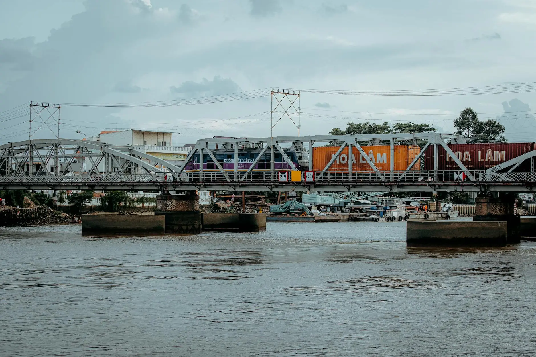 A steel bridge over water with colorful shipping containers and boats nearby.