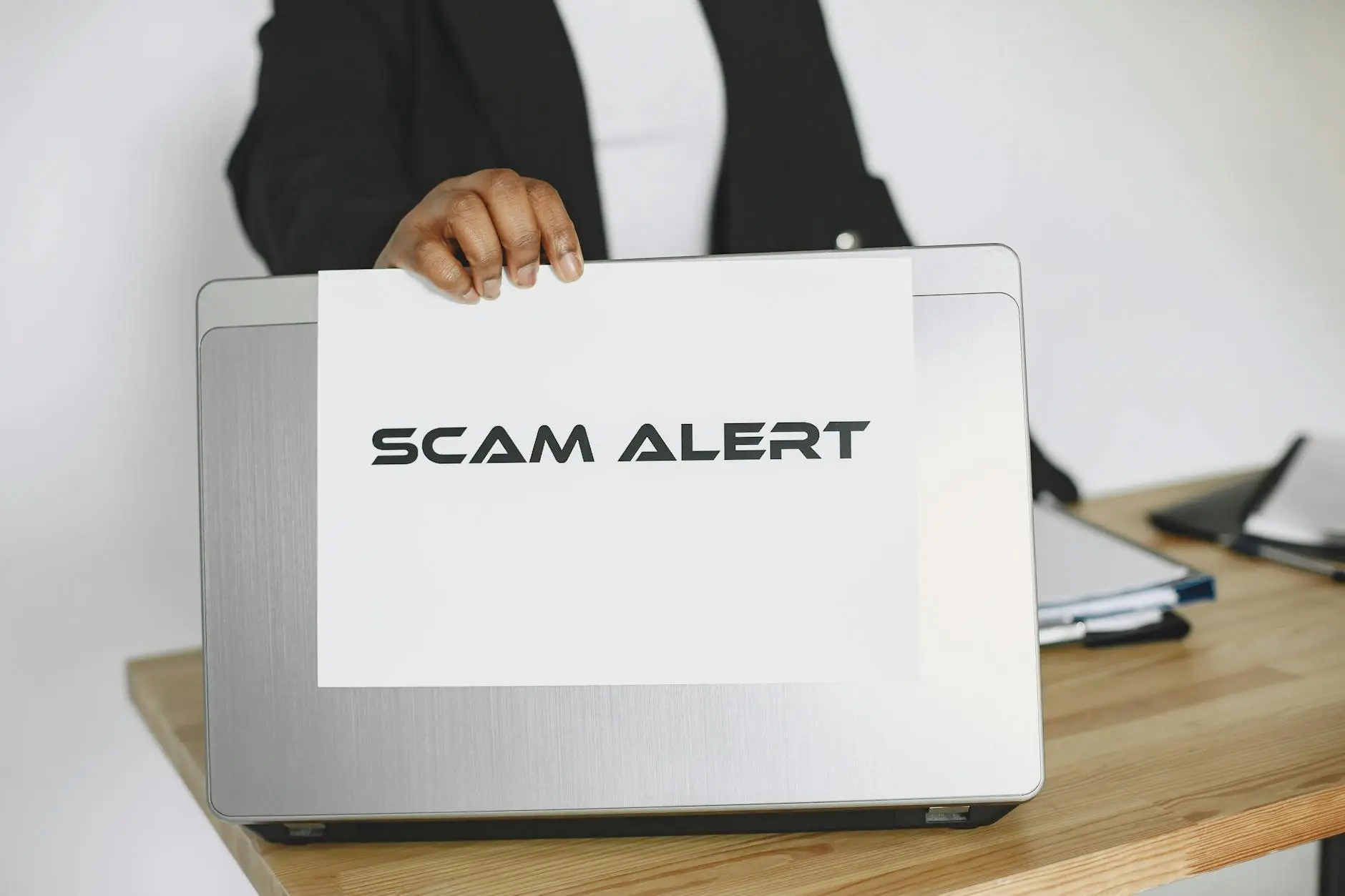 A person holding a 'SCAM ALERT' sign in front of a laptop on a wooden table.
