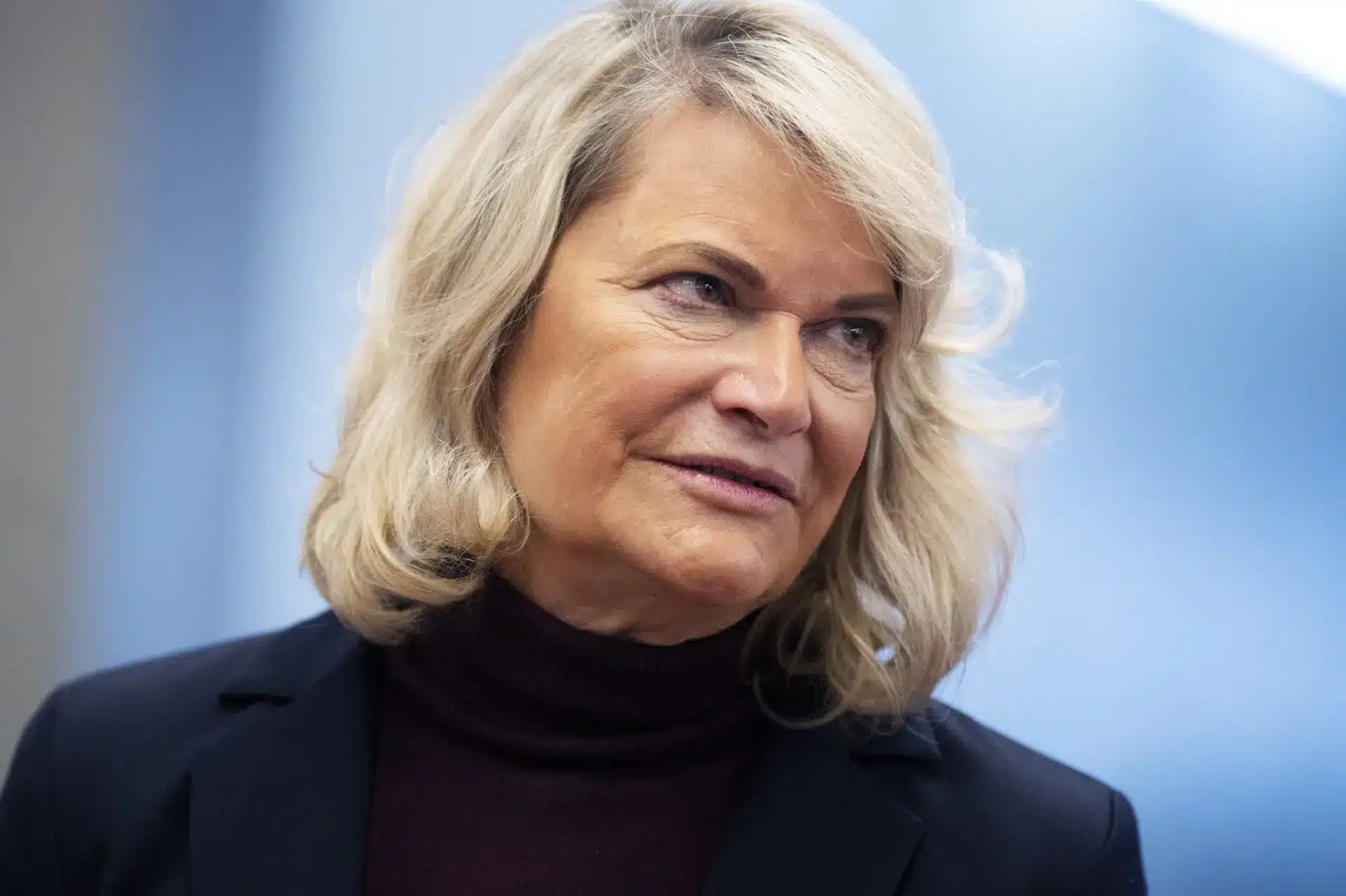 Close-up of a woman with blonde hair, wearing a dark jacket and turtleneck, against a light background.