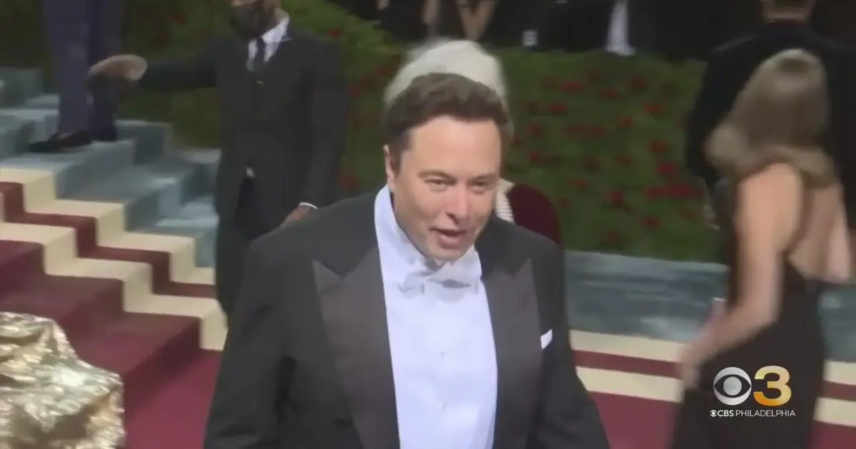 Elon Musk in a tuxedo walking at a red carpet event with blurred crowd in background.