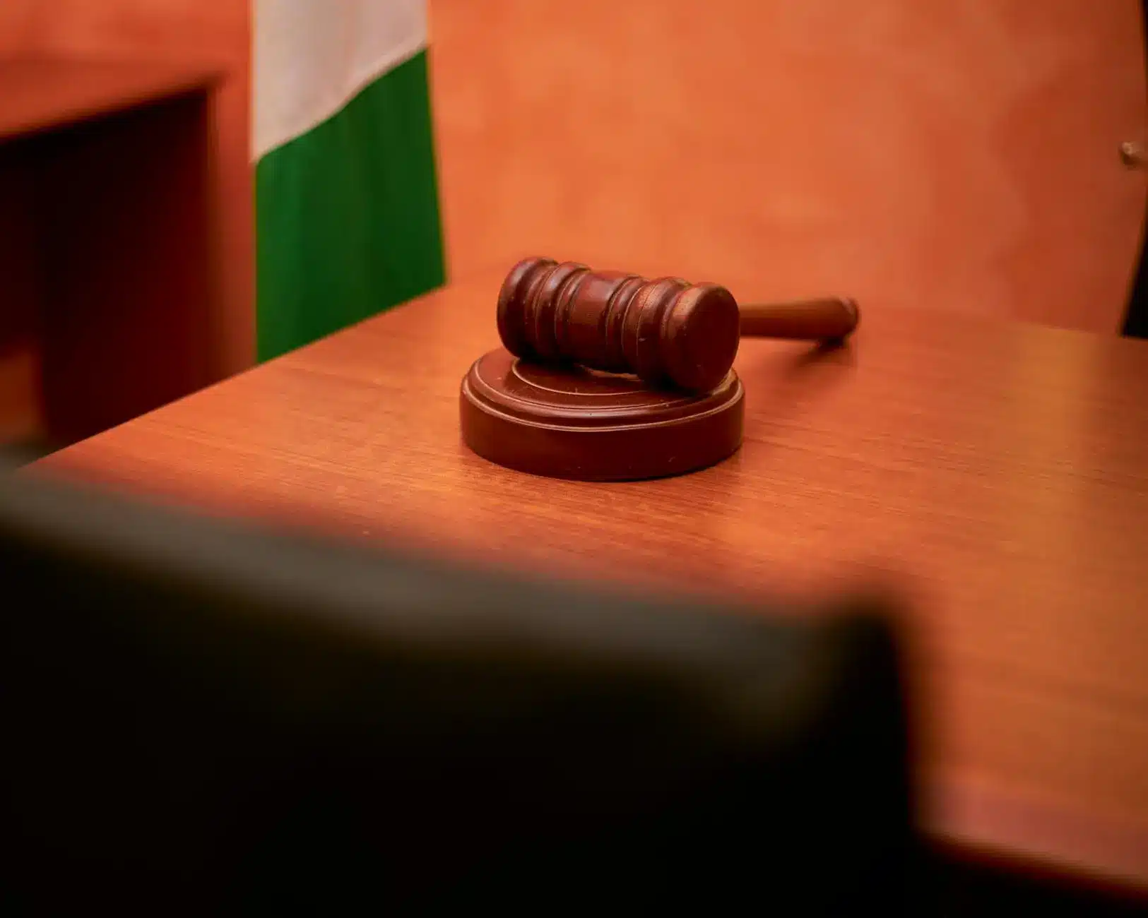 A wooden gavel positioned on a polished desk with a flag in the background.