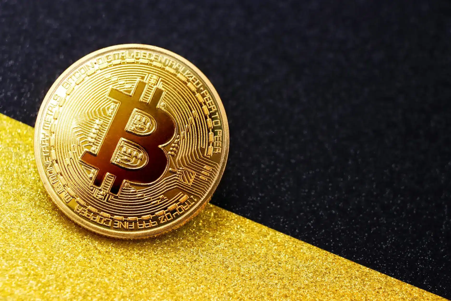 A close-up of a shiny gold Bitcoin placed on a black and gold textured surface.
