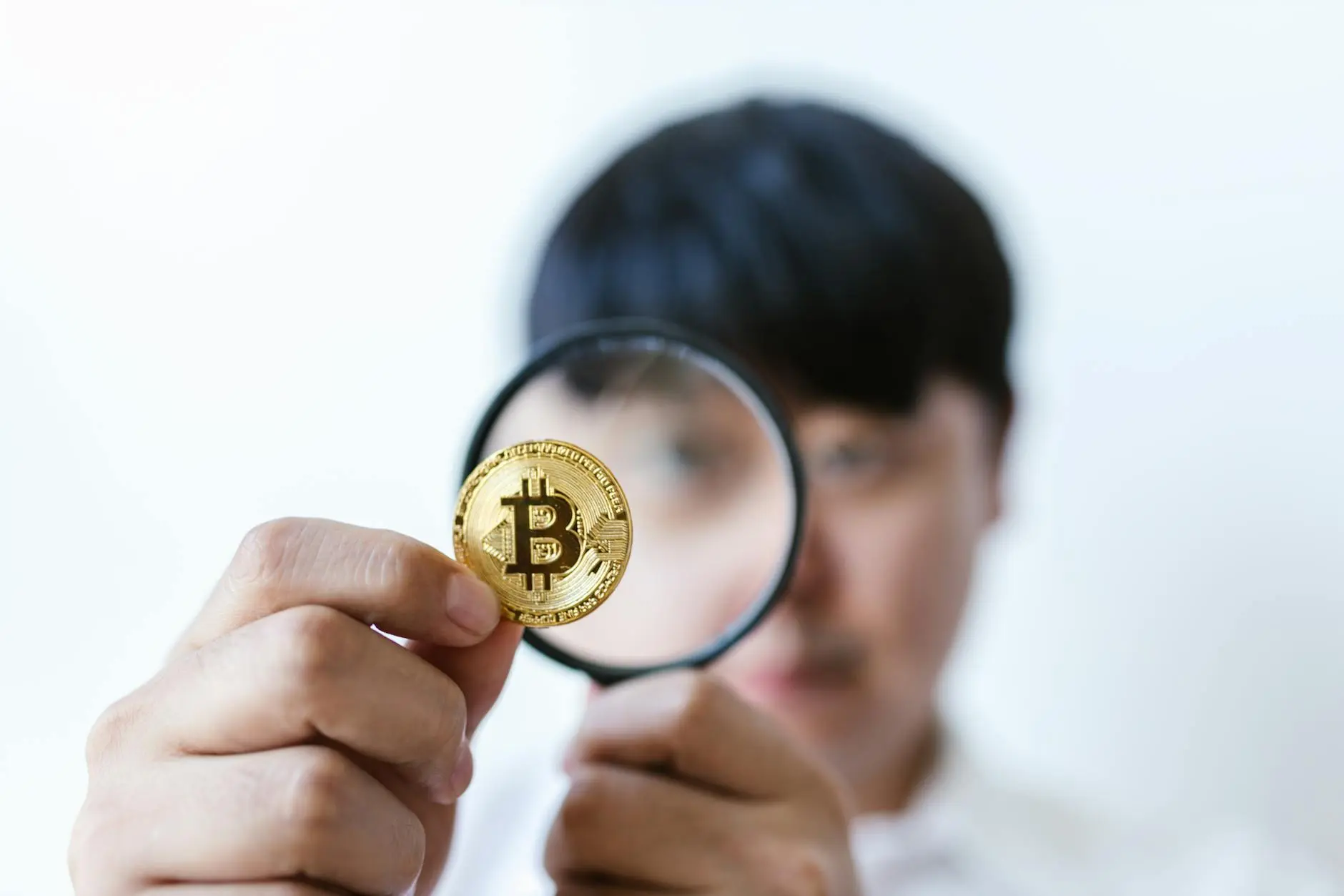 A person examines a Bitcoin coin through a magnifying glass against a light background.