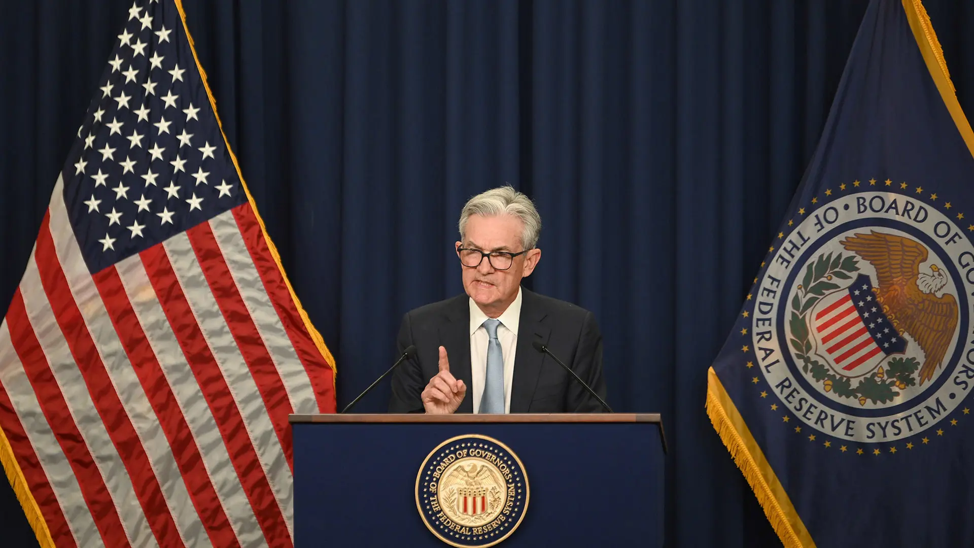Jerome Powell speaking at a podium with the Federal Reserve seal and U.S. flag in the background.