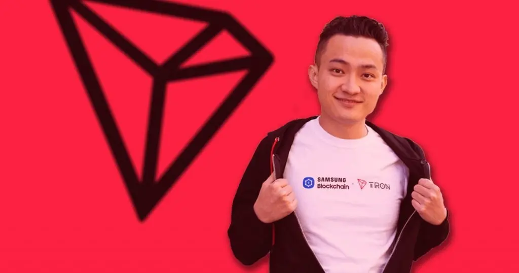 A smiling man in a white t-shirt showing off a crypto logo, against a red background.