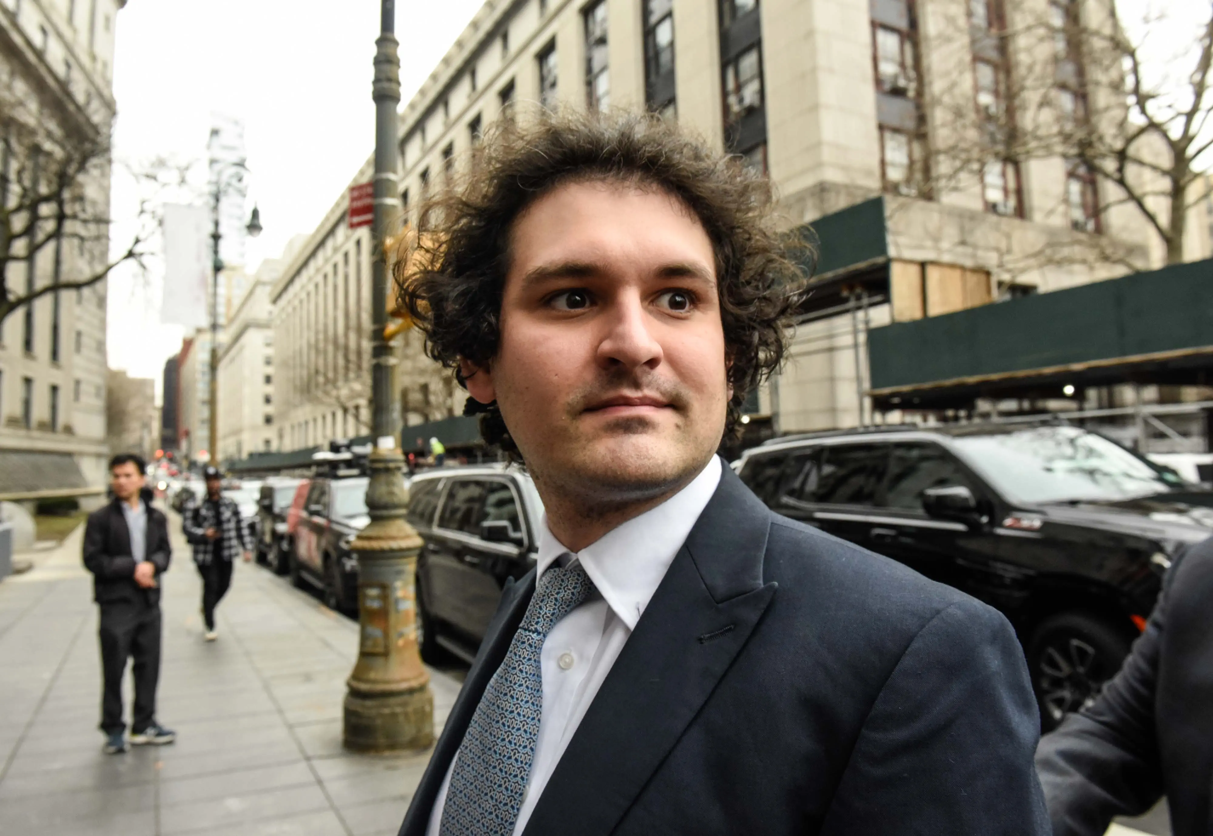 Sam Bankman-Fried walking on the street, wearing a suit with buildings and cars in the background.