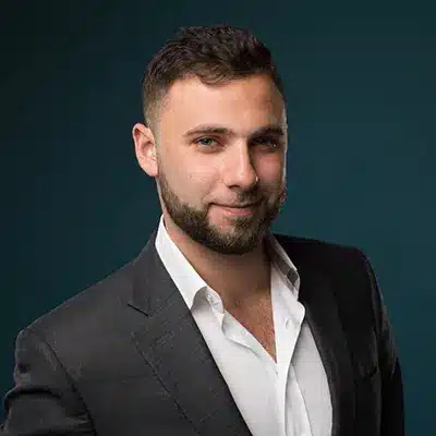 A young man in a blazer smiling against a dark blue background.