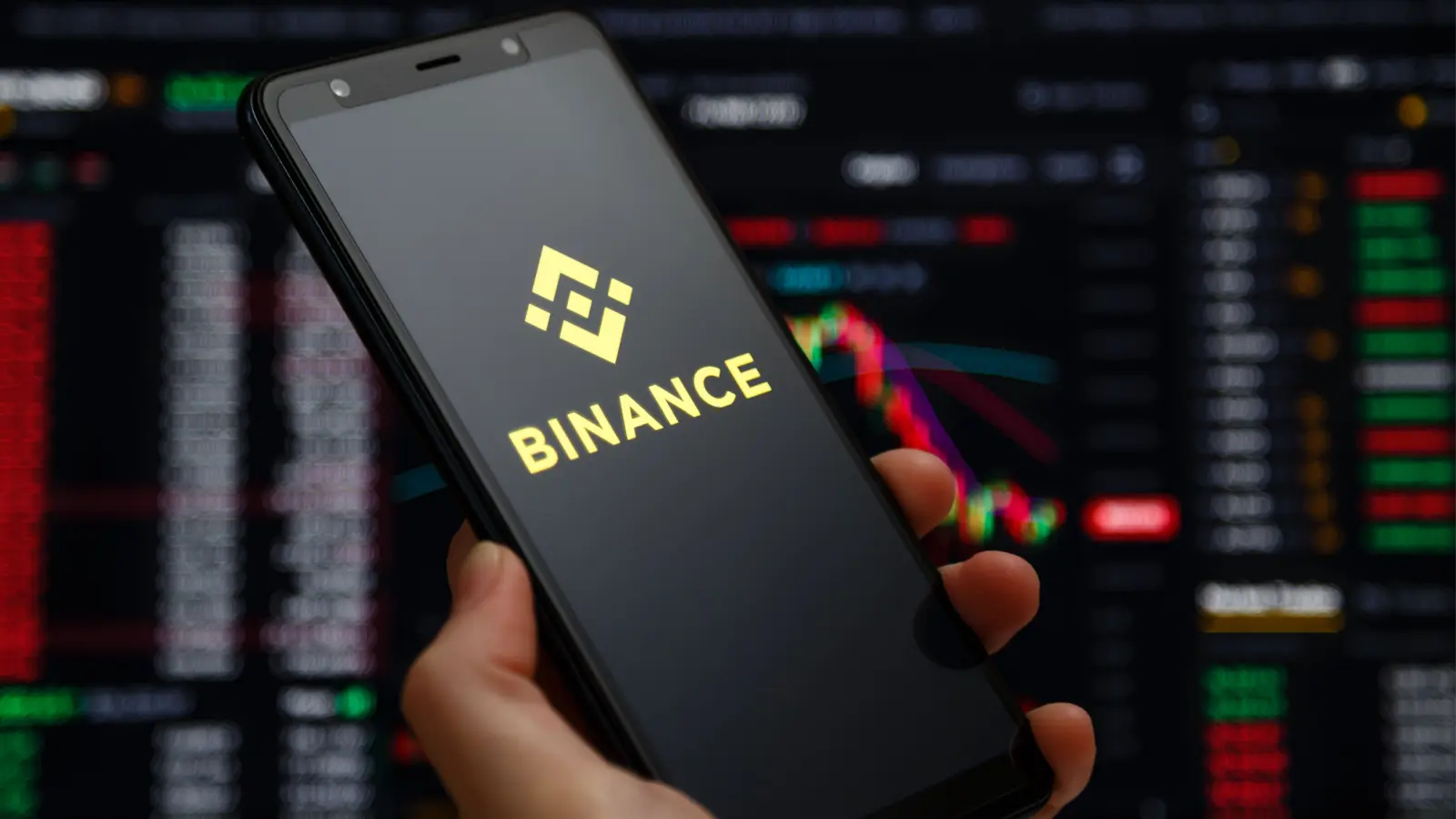 Hand holding a smartphone displaying the Binance logo with charts in the background.