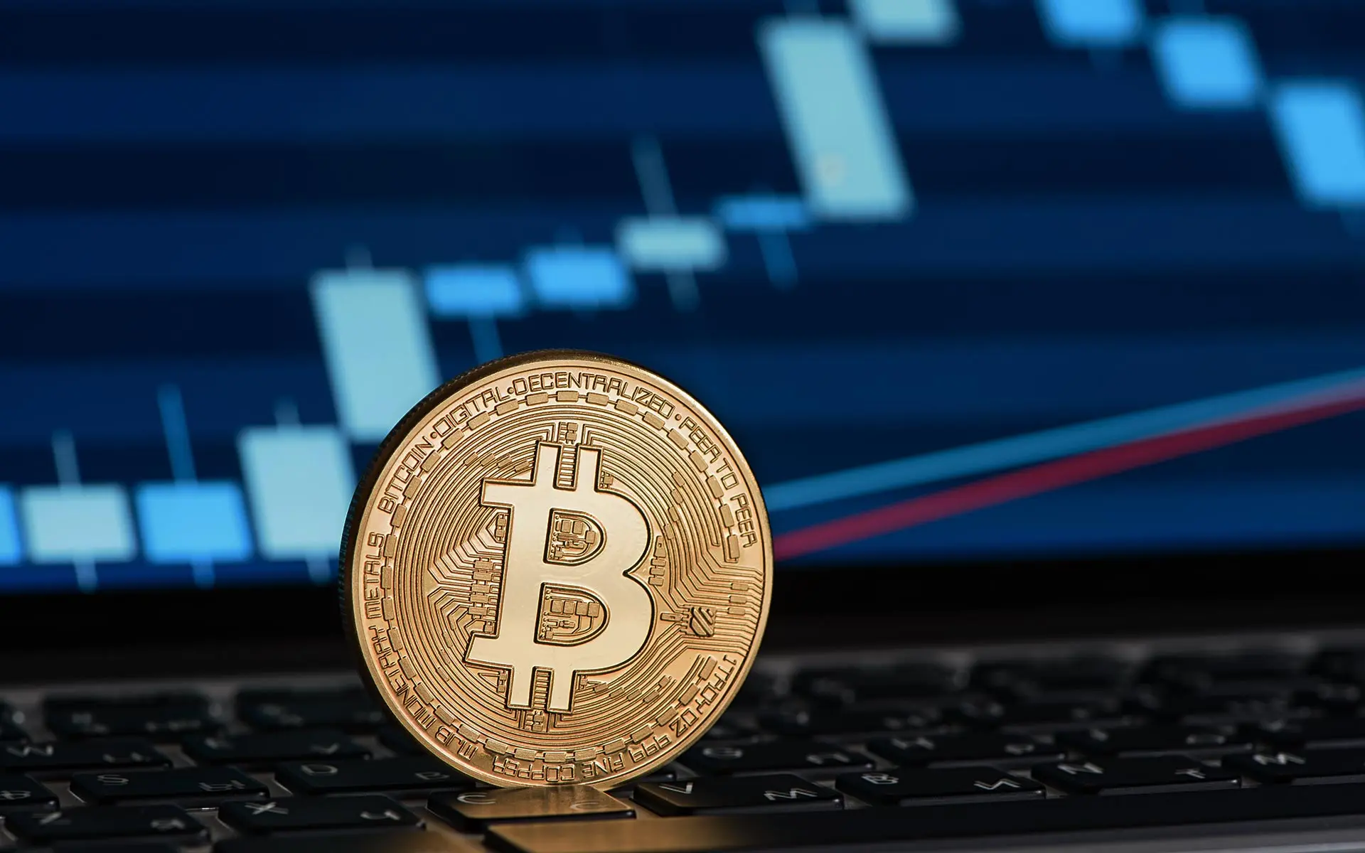 Close-up of a Bitcoin coin on a laptop keyboard with a trading chart in the background.