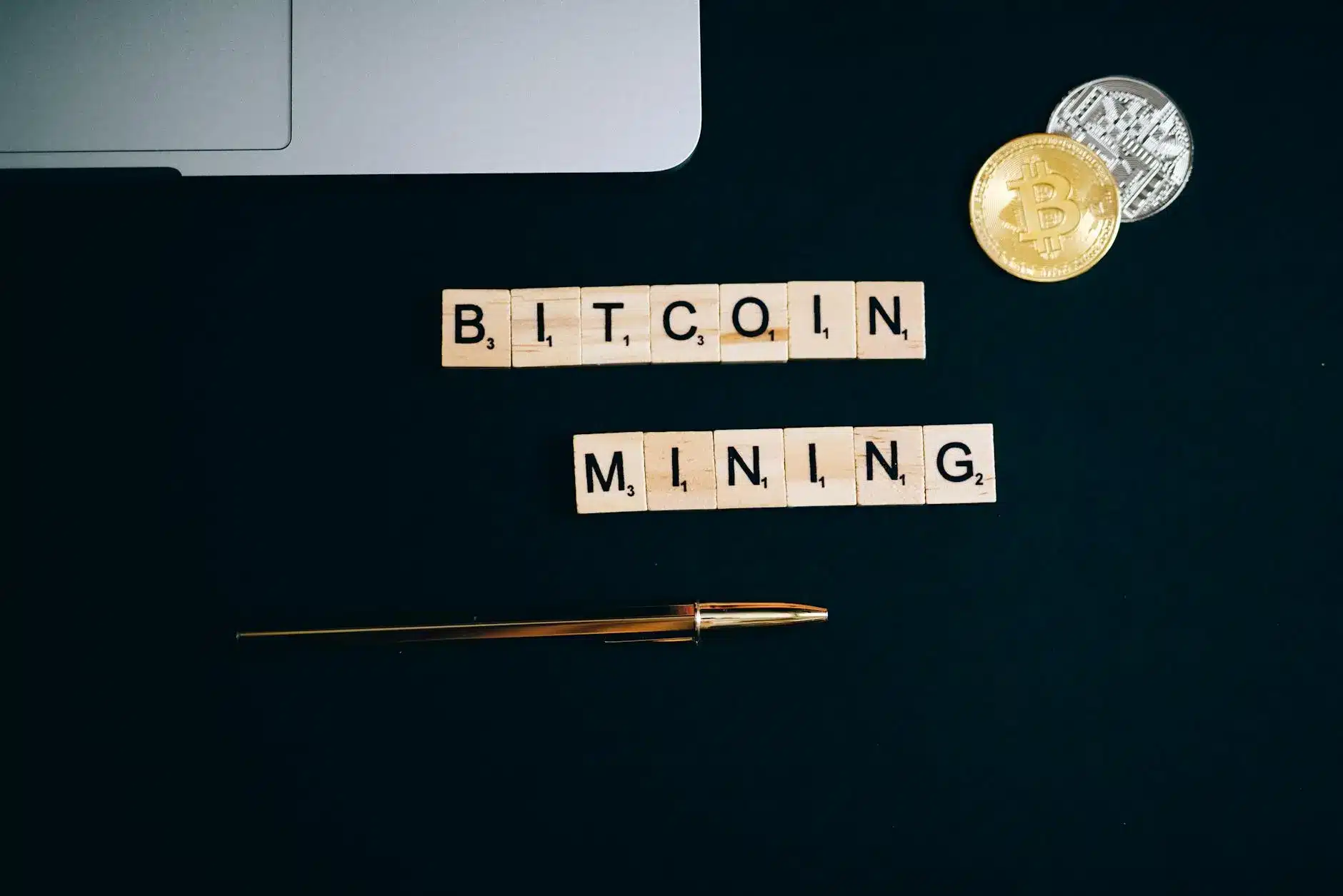 Scrabble pieces spelling 'Bitcoin Mining' with a pen and coins on a dark background.