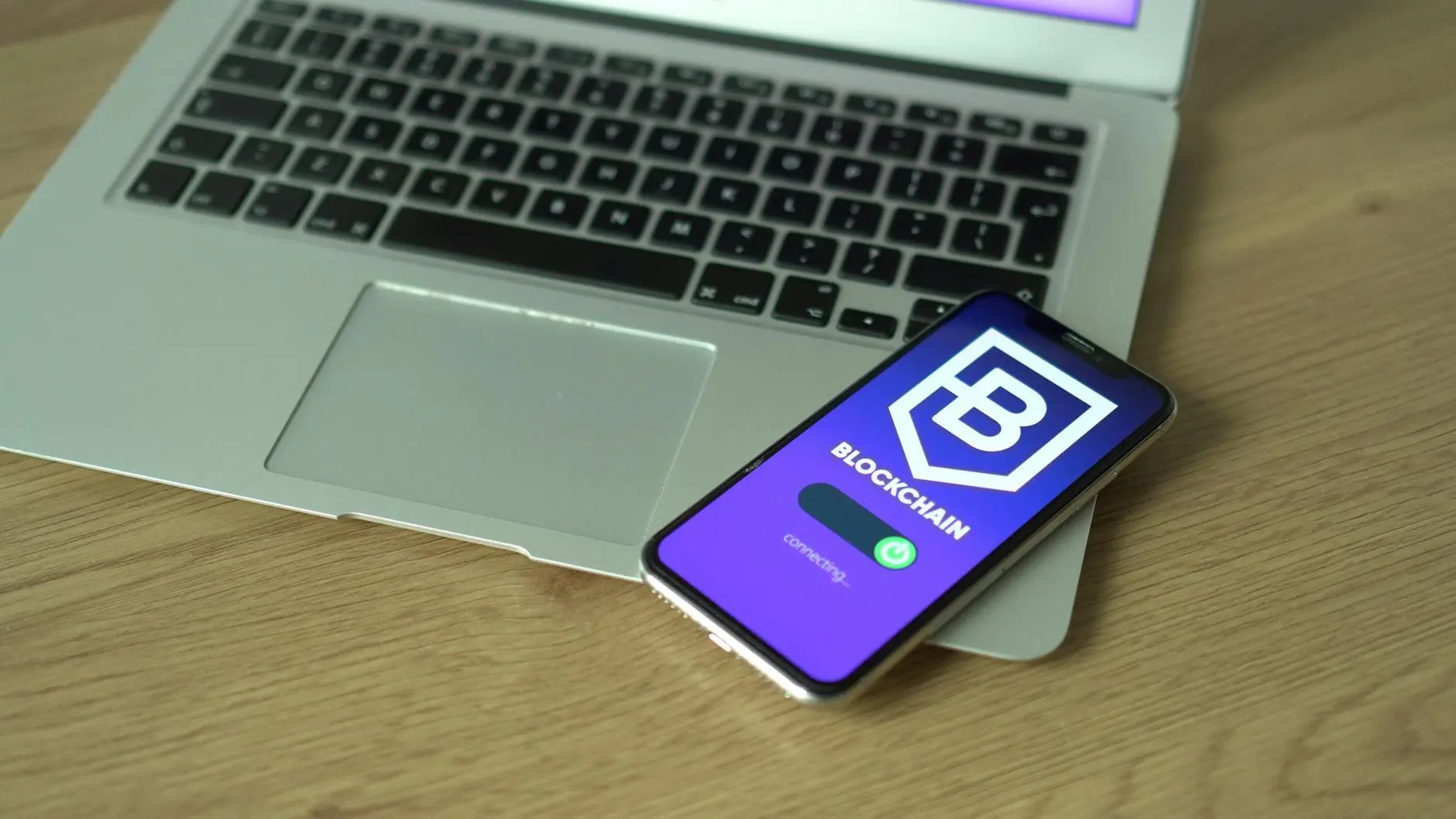 Mobile phone displaying blockchain app next to a laptop on wooden desk.