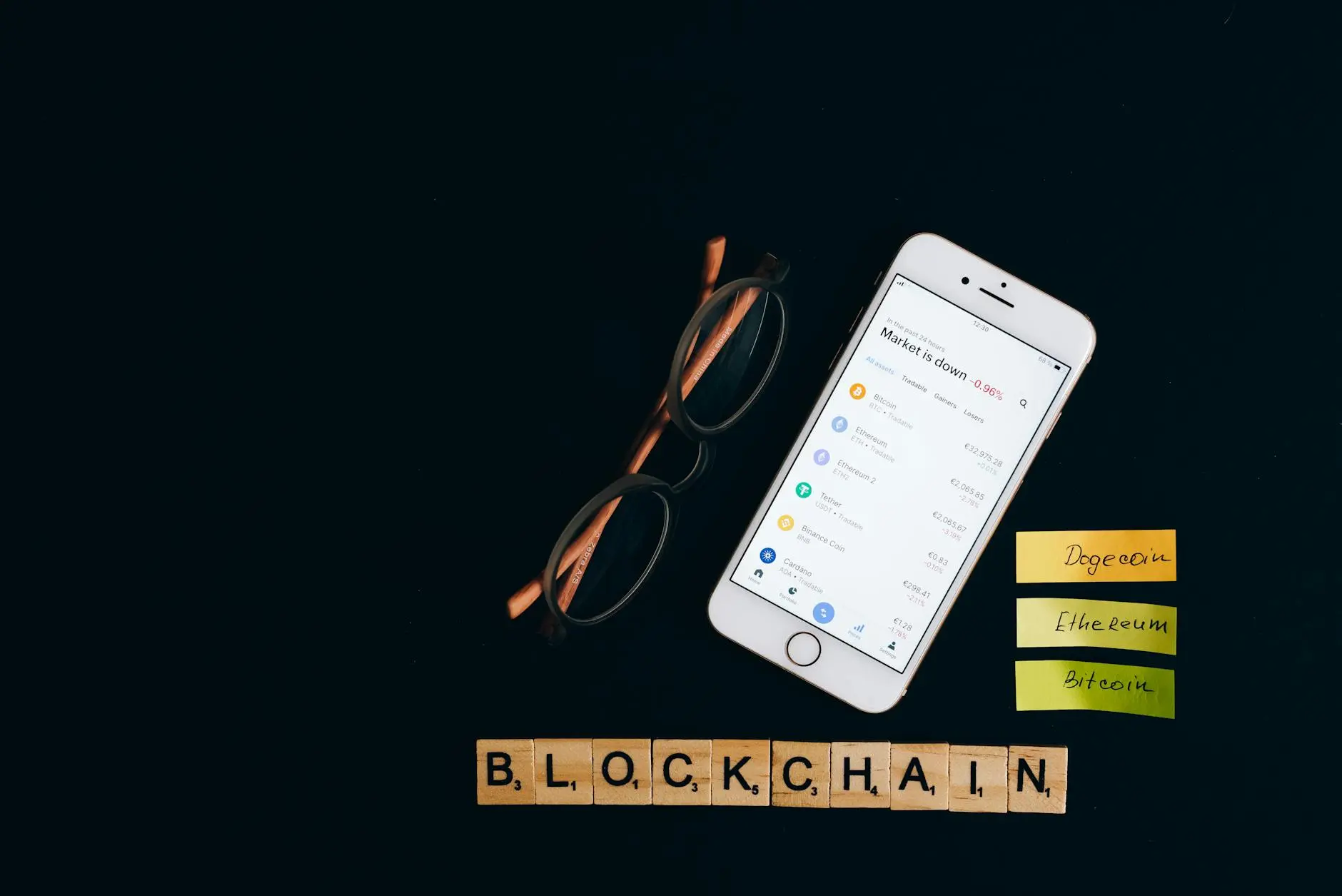 A smartphone displaying cryptocurrency values next to glasses and wooden letter blocks spelling 'BLOCKCHAIN'.