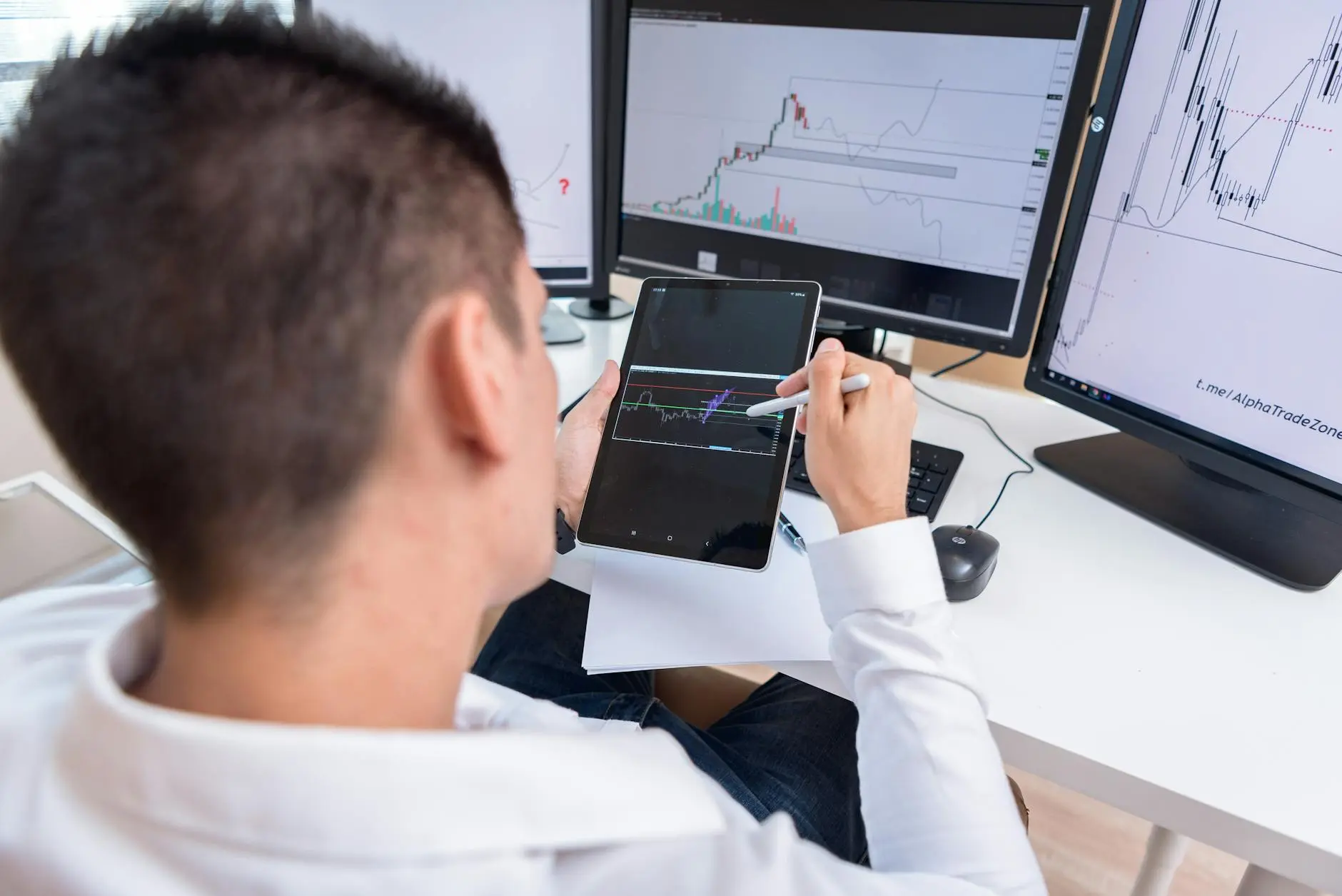Person analyzing crypto charts on a tablet with dual monitors at a workstation.