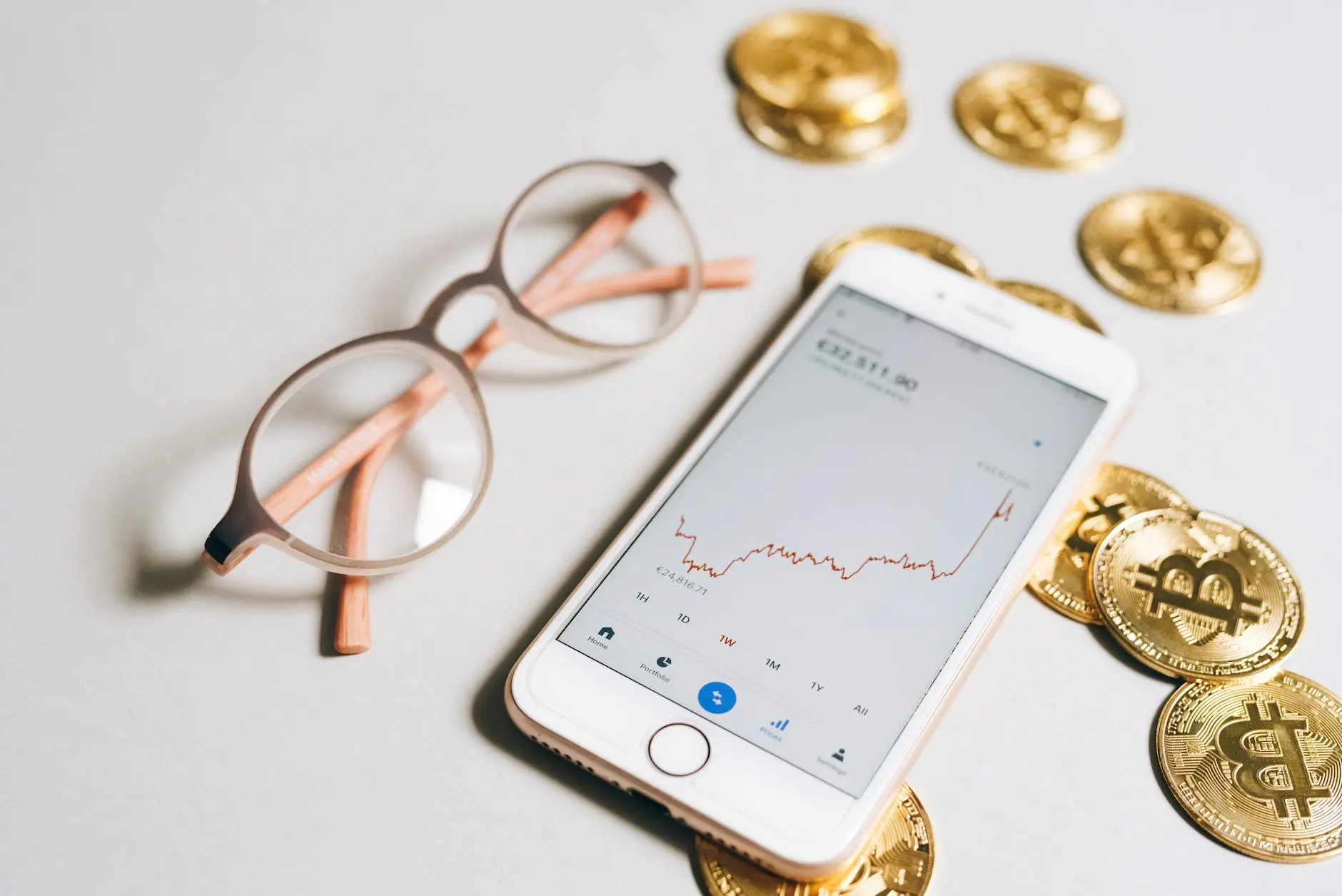 Smartphone displaying cryptocurrency chart next to golden Bitcoin coins and glasses.