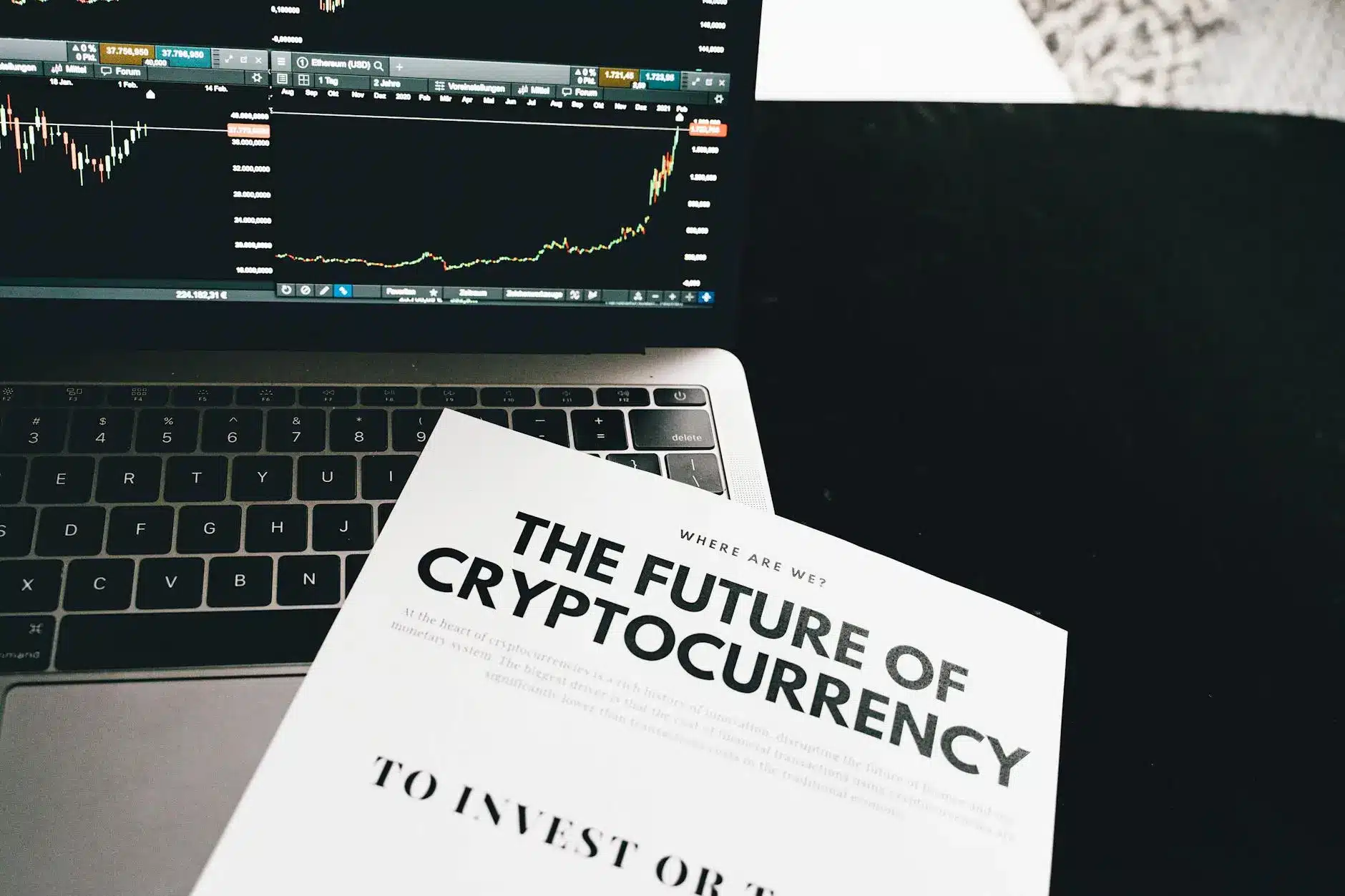 A laptop displaying cryptocurrency charts next to a book titled 'The Future of Cryptocurrency'.