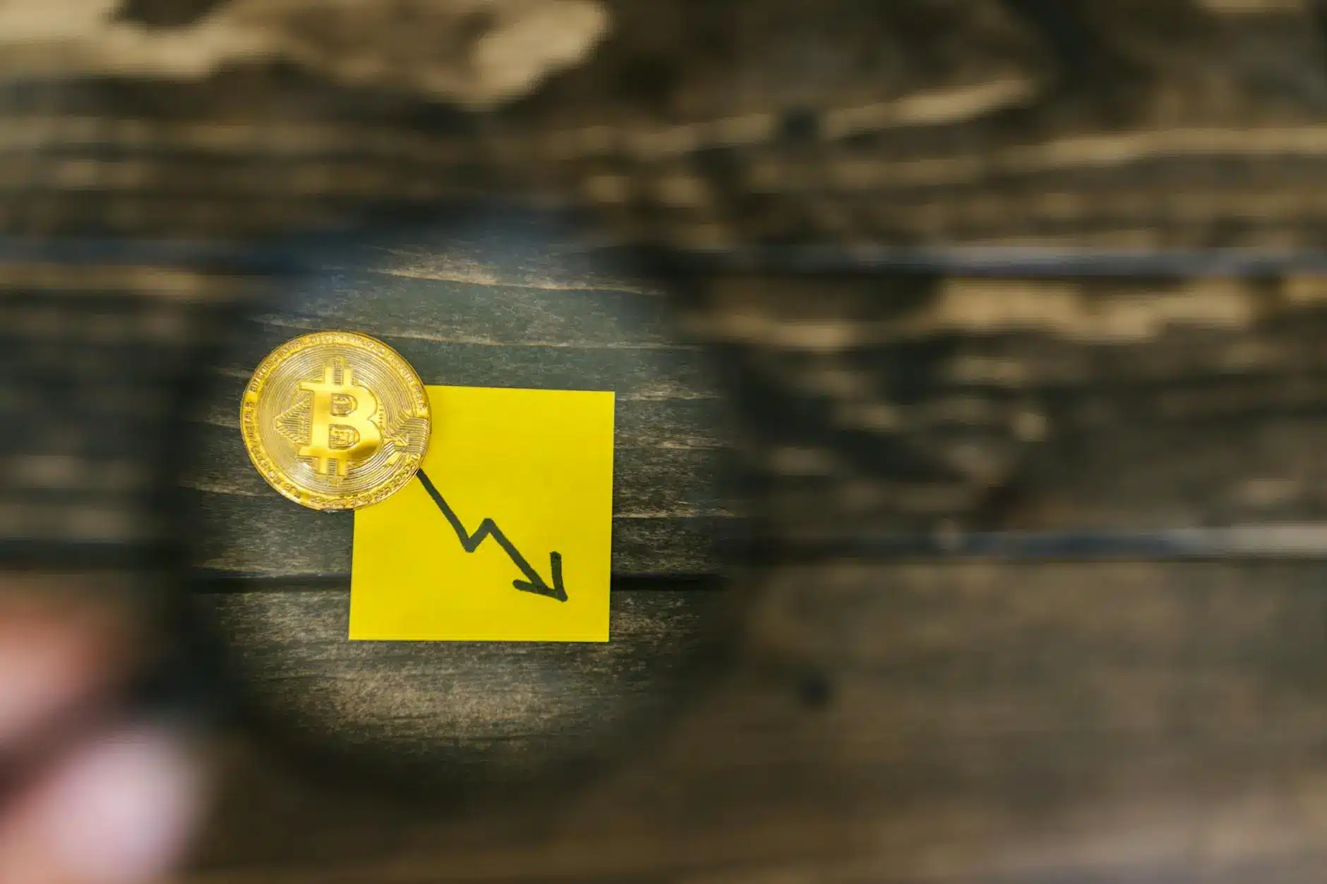 A Bitcoin coin placed on a yellow sticky note with a downward trend graph illustration.