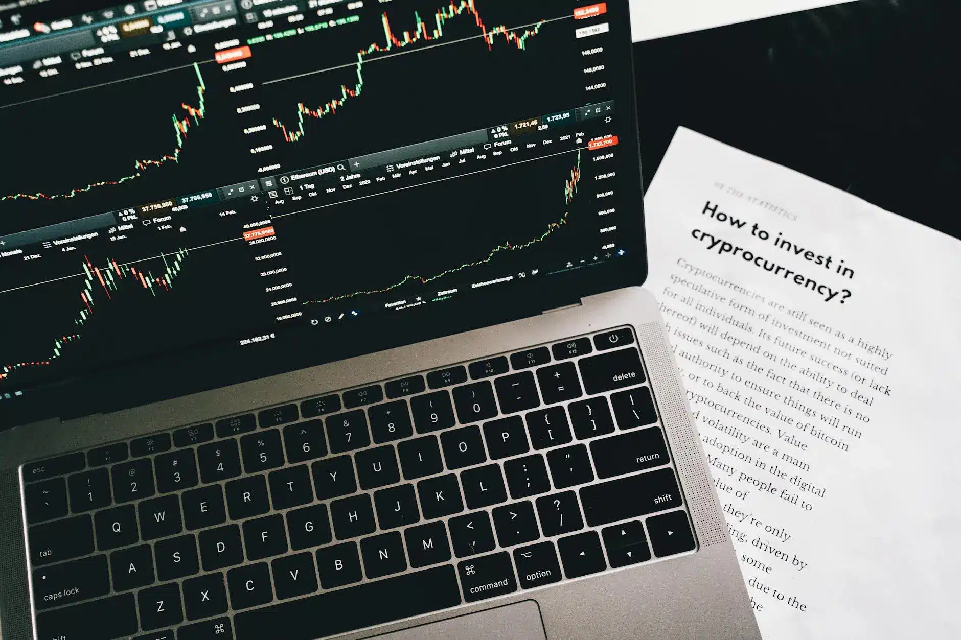 A laptop displaying cryptocurrency trading charts and a paper about investing in cryptocurrency.