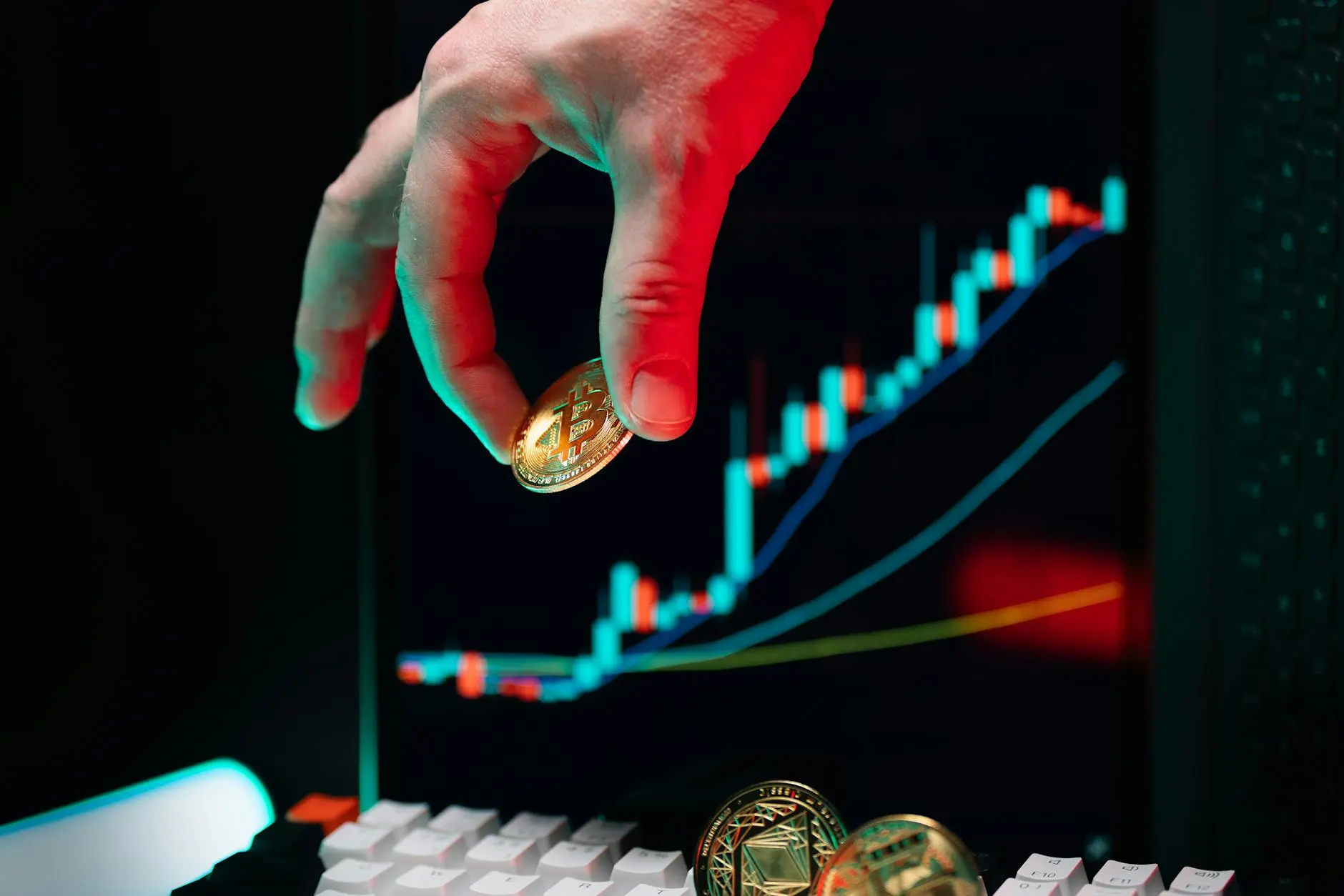 A hand holding a Bitcoin above a computer keyboard and a cryptocurrency market chart.