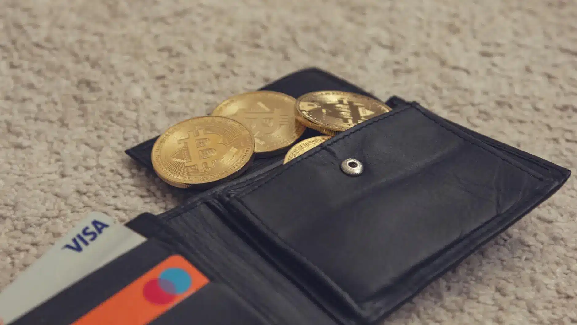 A black wallet containing gold coins with Bitcoin symbols and credit cards.