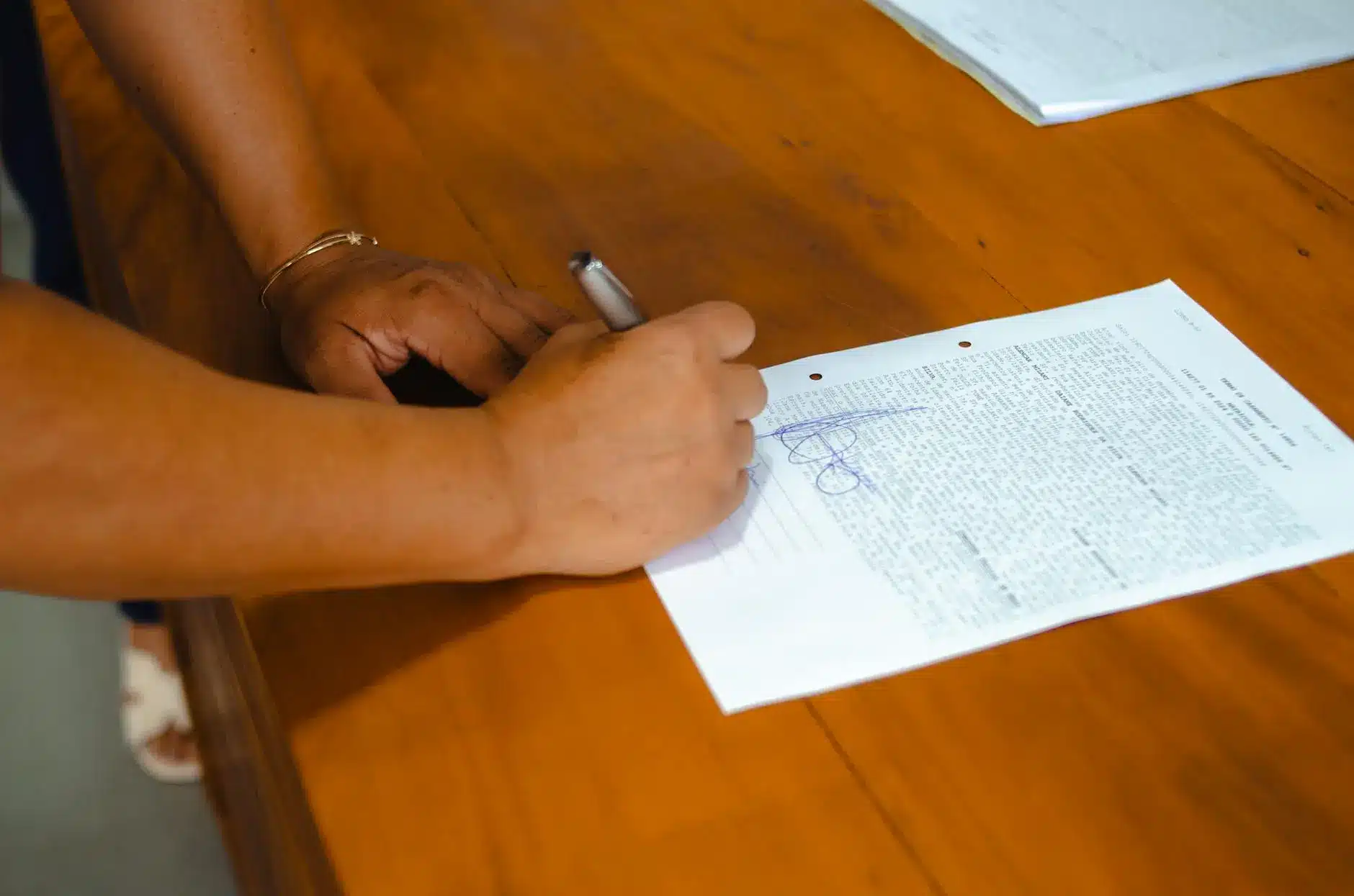 A person writing on a sheet of paper with printed text on a wooden table.