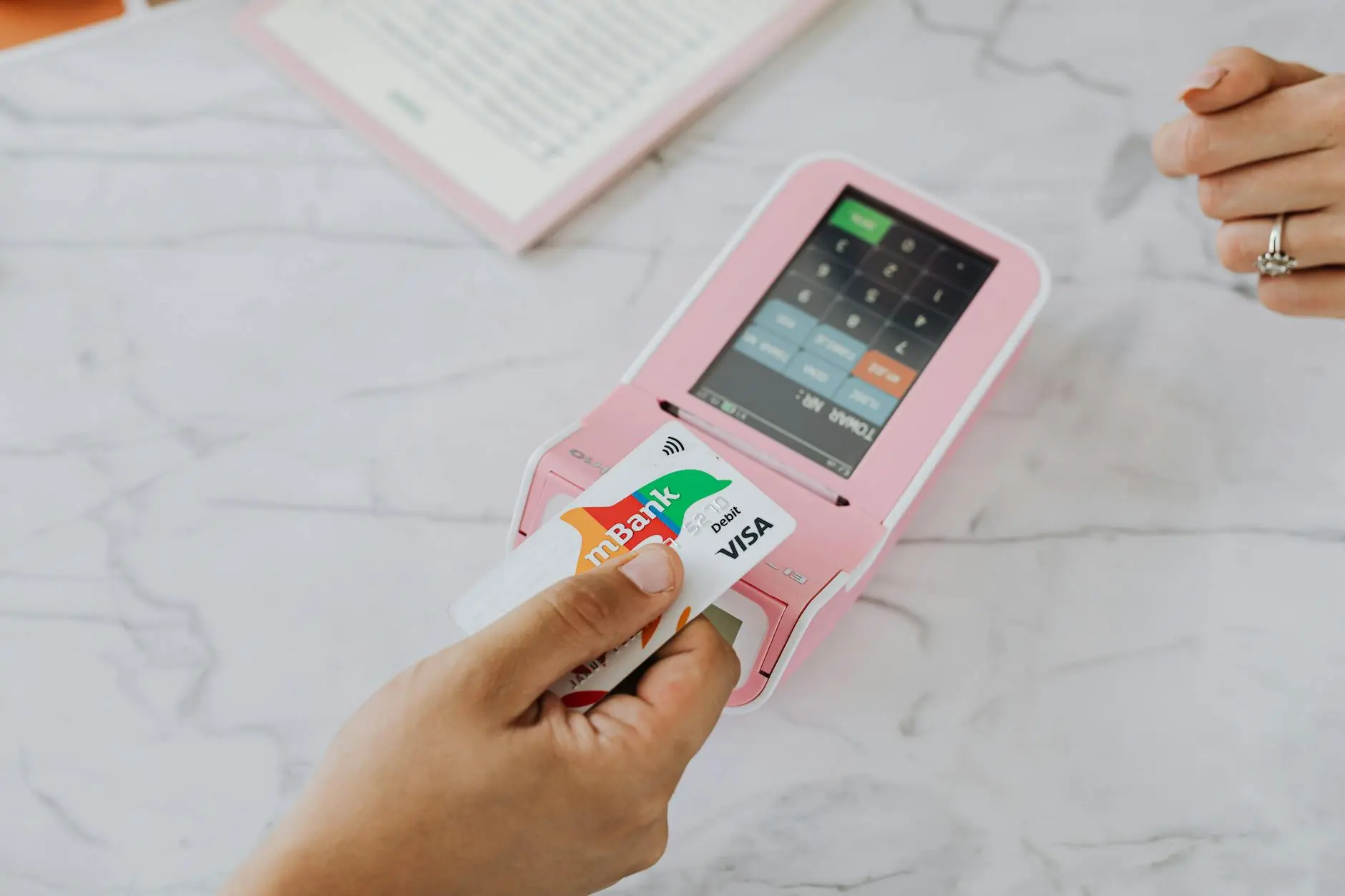 A hand using a Visa debit card on a pink payment terminal on a marble surface.