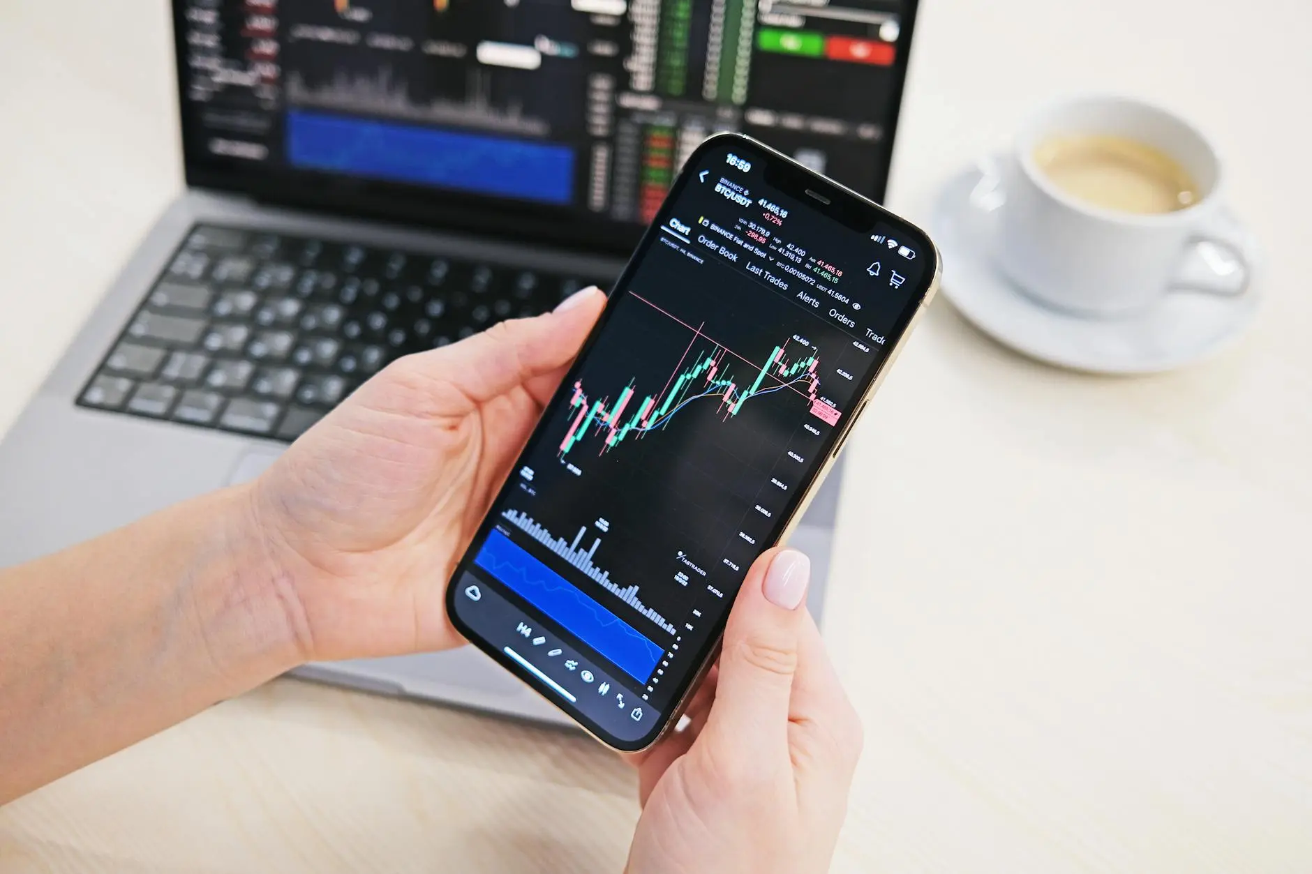 A person holding a smartphone displaying a trading chart, with a laptop in the background.