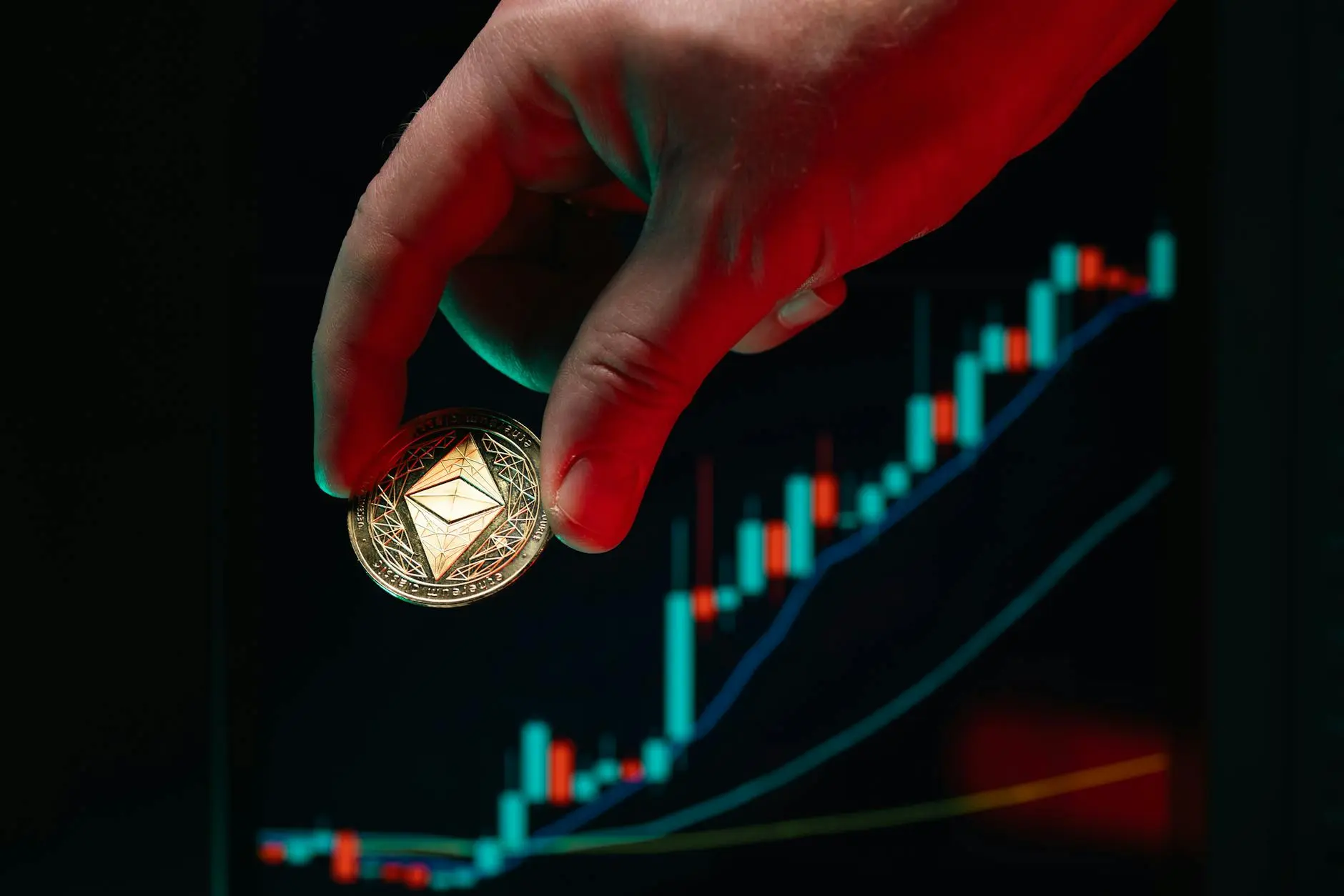A hand holding a gold Ethereum coin in front of a colorful trading chart.