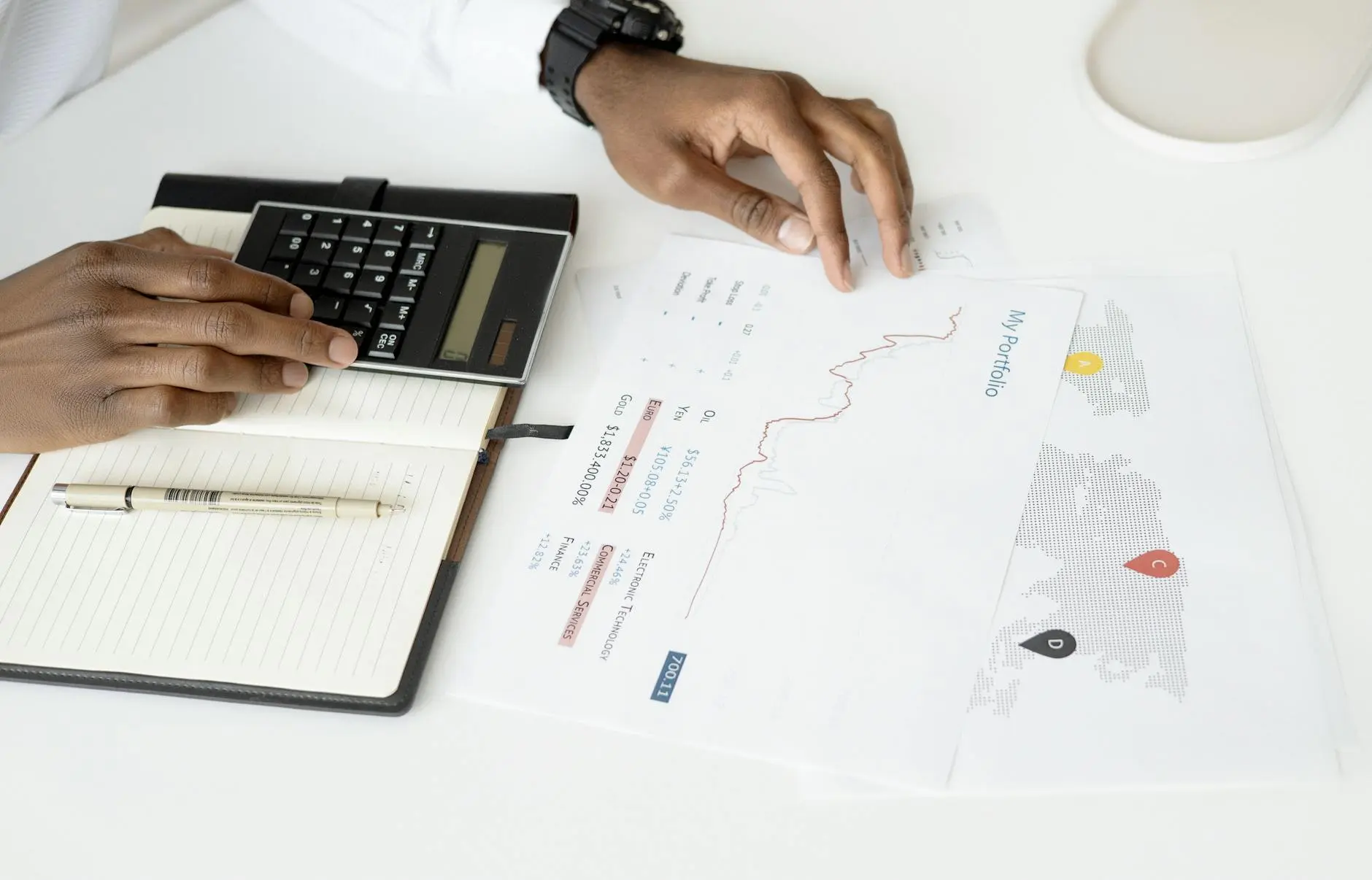 A person using a calculator beside portfolio documents and a notebook on a desk.