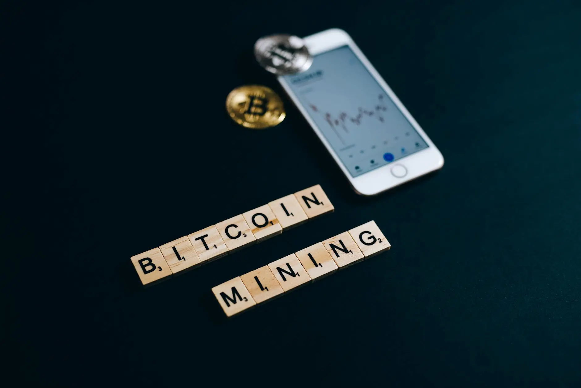 Scrabble tiles spelling 'BITCOIN MINING' beside a smartphone displaying a Bitcoin chart.