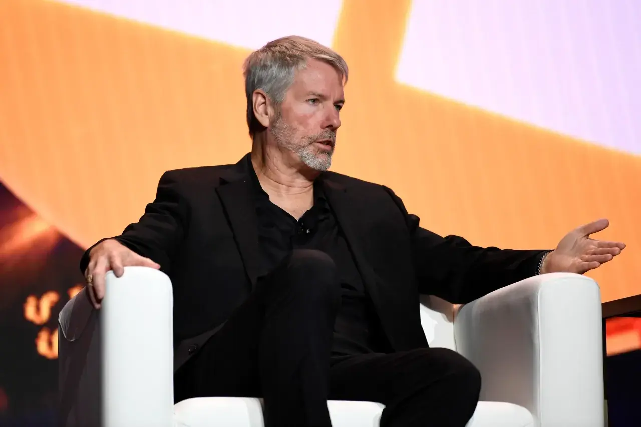 Michael Saylor speaking at a Bitcoin conference, sitting on a white chair.
