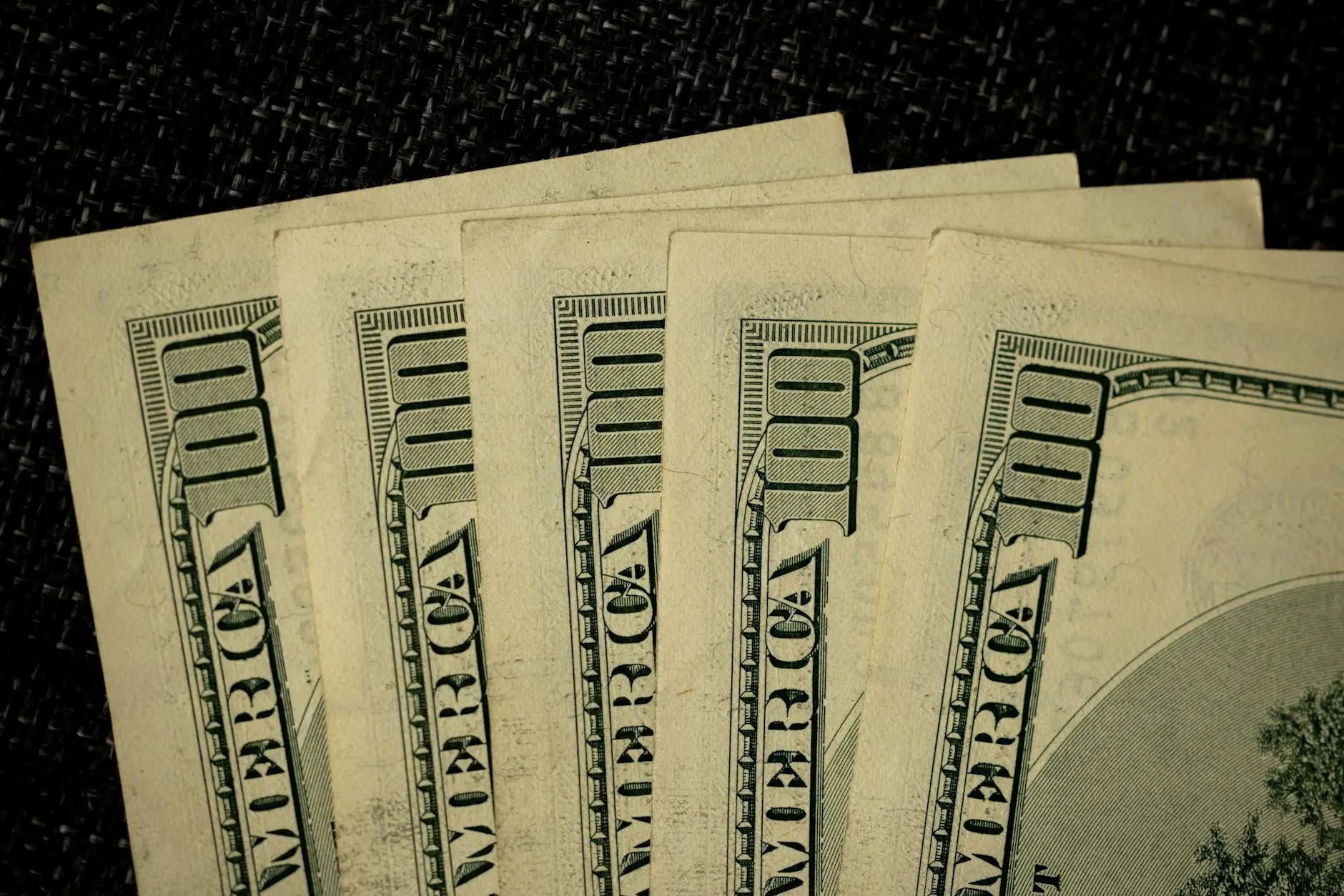 Close-up of several stacked hundred-dollar bills on a dark textured surface.