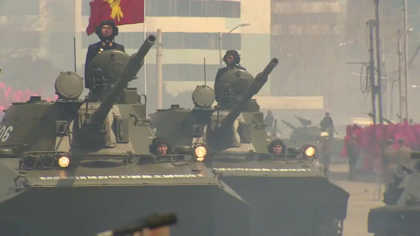 North Korean military tanks advance in a parade, with soldiers and flags around.