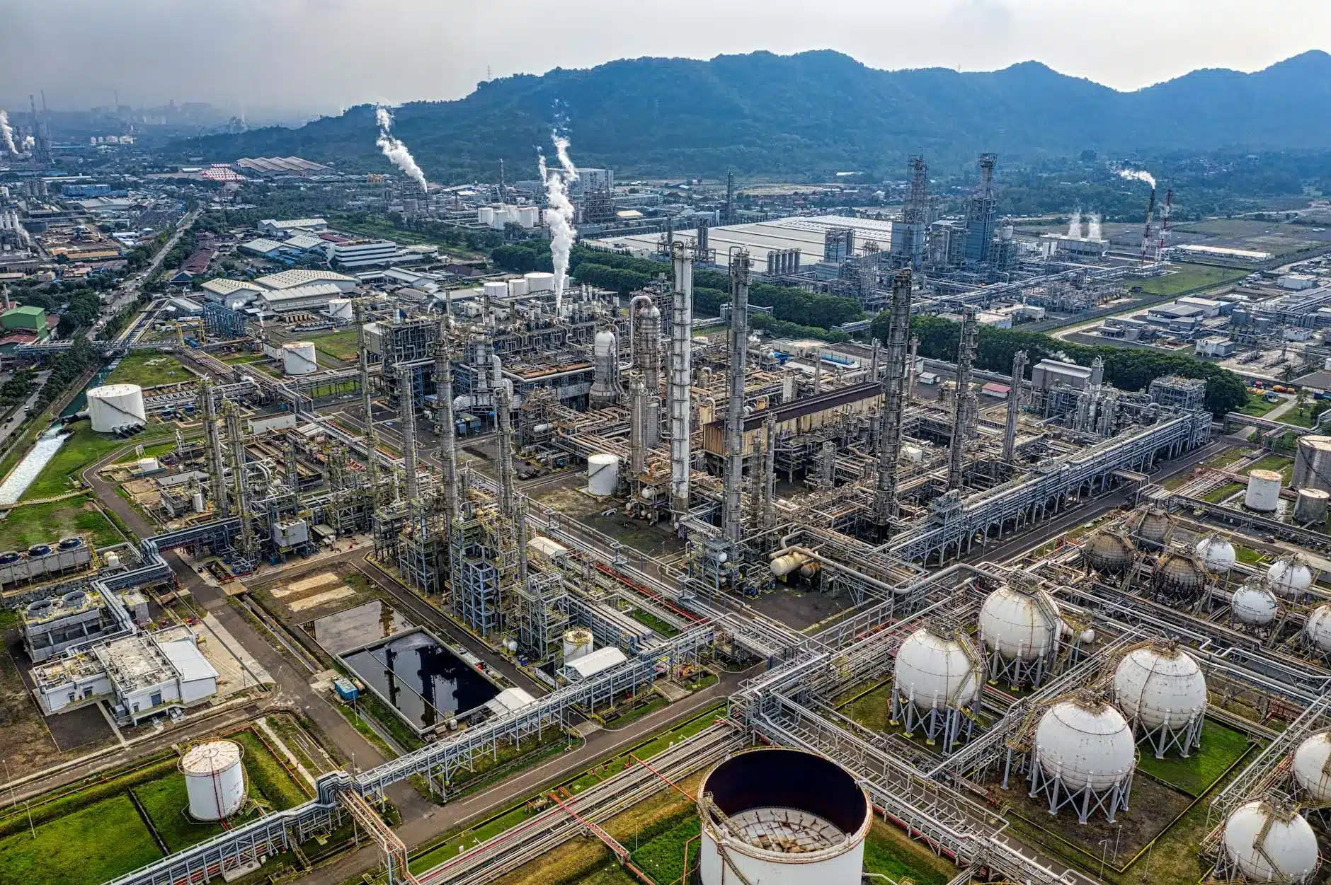 Aerial view of an oil refinery with pipelines and storage tanks under a cloudy sky.