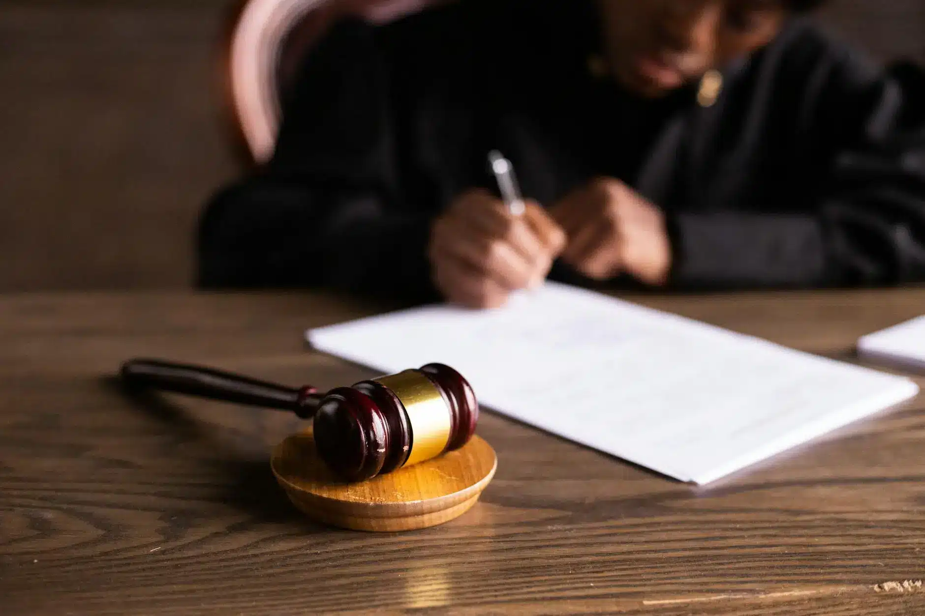 A gavel on a wooden base with a person writing on legal documents.