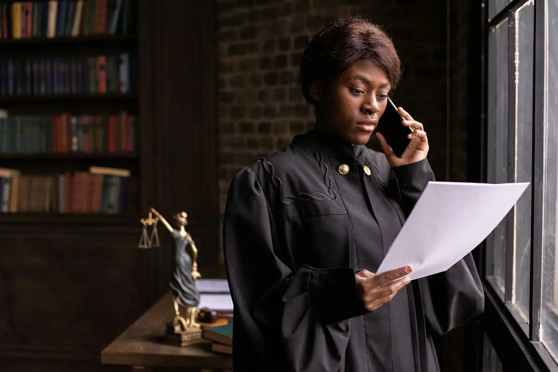 A woman in judicial robes holds a document while talking on a phone in a library setting.