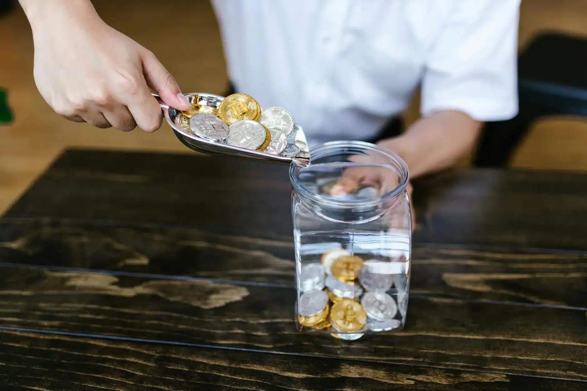 A person adding various cryptocurrency coins into a clear glass jar.