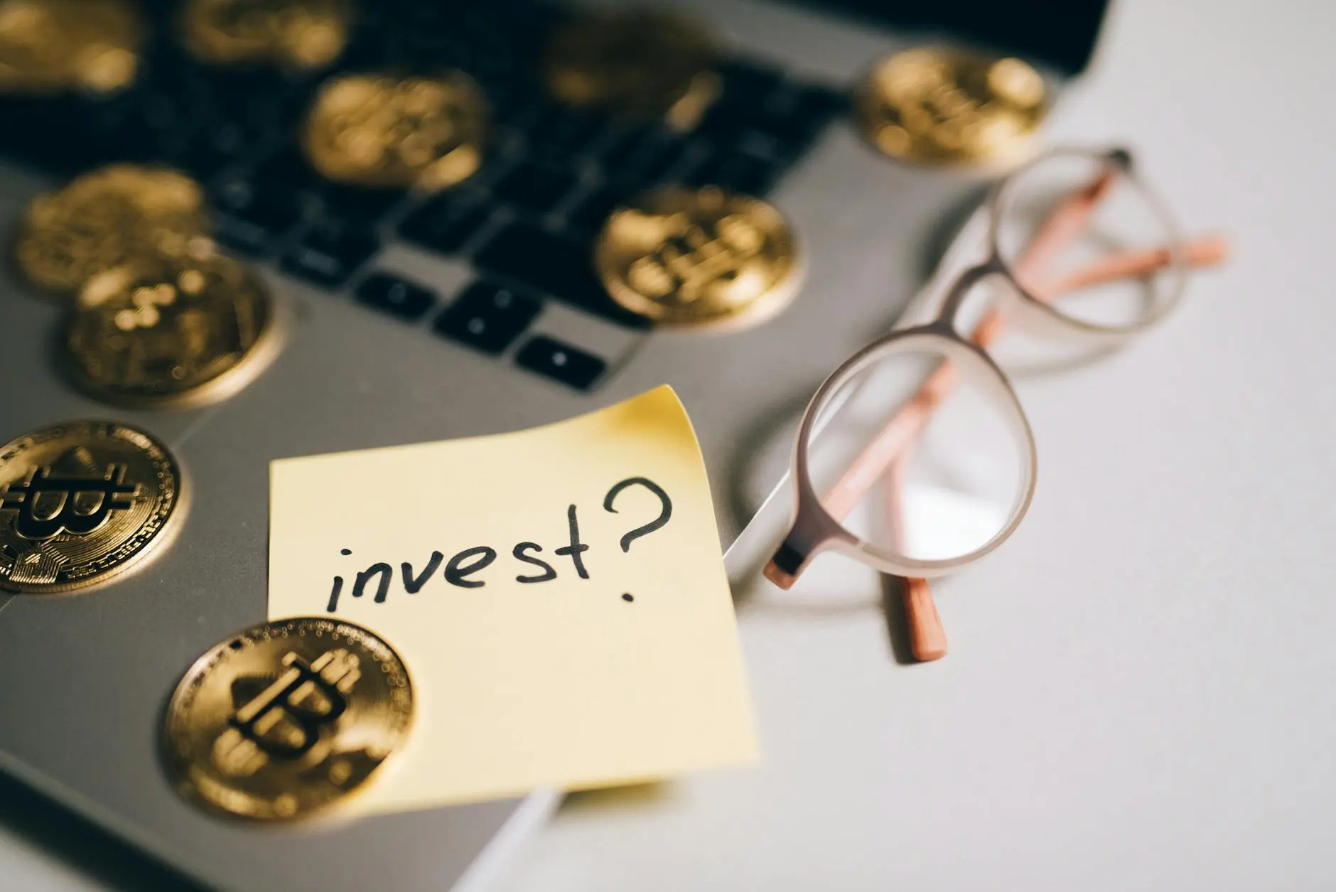 A laptop with gold cryptocurrency coins and a sticky note saying 'invest?'